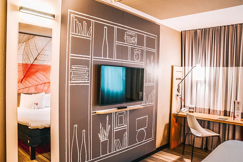 Hotelzimmer mit wandmontiertem Fernseher in einer grafisch bemalten Wand, Schreibtisch mit Stuhl, Vorhänge und Bett im Spiegel.