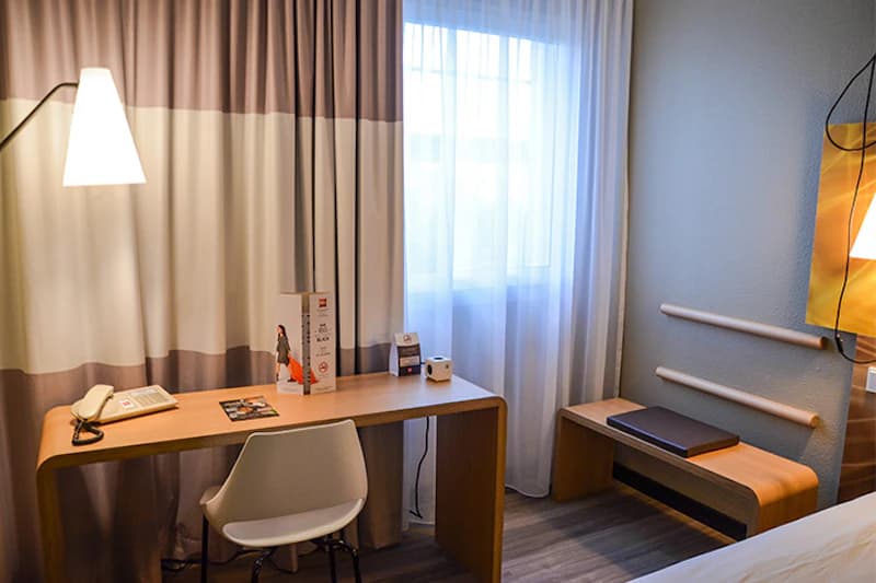 Schreibtisch mit Stuhl, Telefon, Broschüren, stehender Lampe, Fenster mit Vorhängen und Sitzbank.