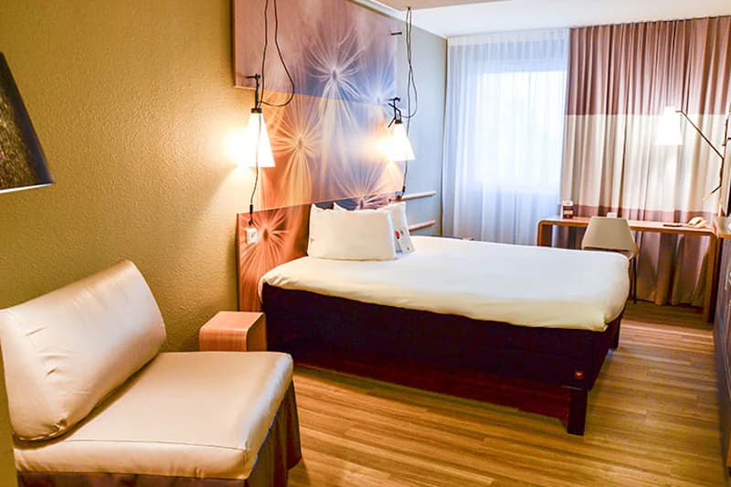 Hotelzimmer mit Doppelbett, dekorativer Kopfwand, hängenden Nachttischlampen, Sessel und Schreibtisch am Fenster.