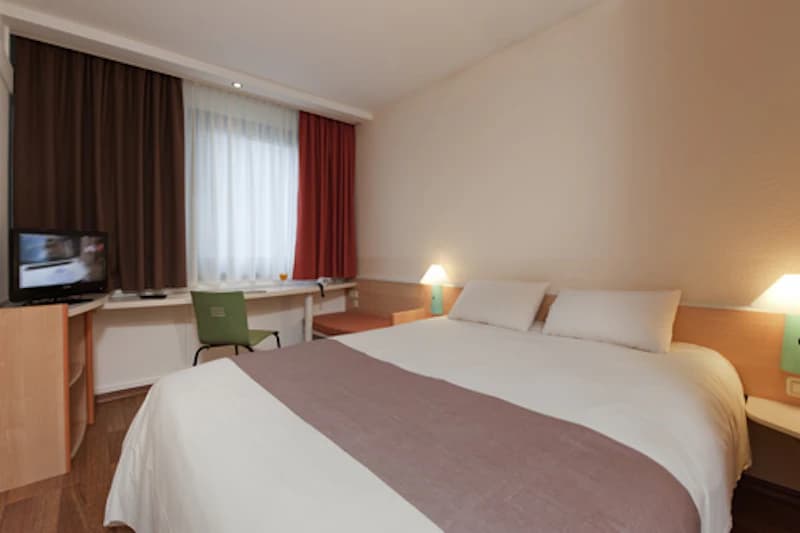 Hotelzimmer mit Doppelbett, Nachttischen, zwei Lampen, Schreibtisch, grünem Stuhl und Fernseher vor einem Fenster.