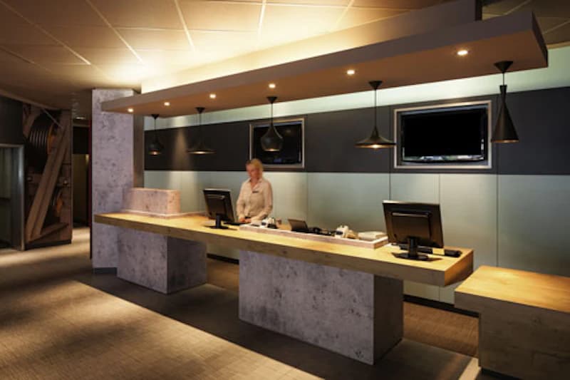 Rezeptionstresen mit Rezeptionistin, zwei Monitoren und Hängeleuchten in moderner Hotellobby.