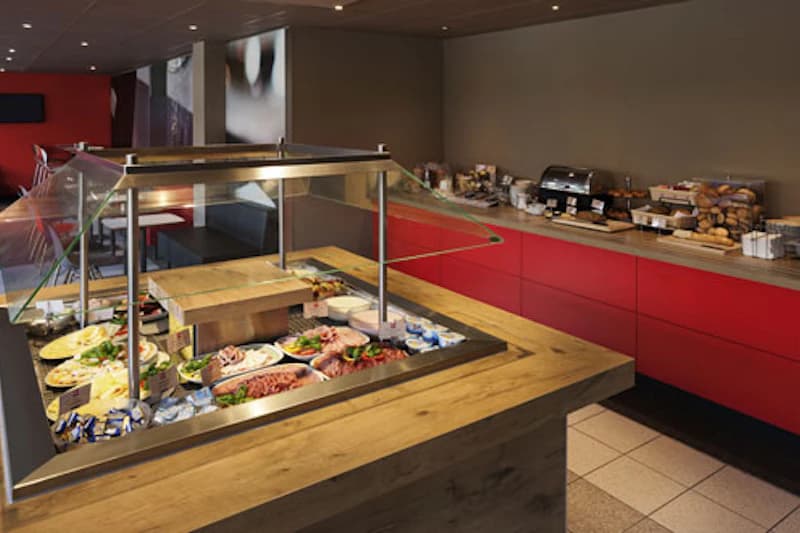 Buffetinsel mit Aufschnitt, Käse und Beilagen, Glasabdeckung; rote Schrankzeile mit Toaster und Brötchen im Hintergrund.
