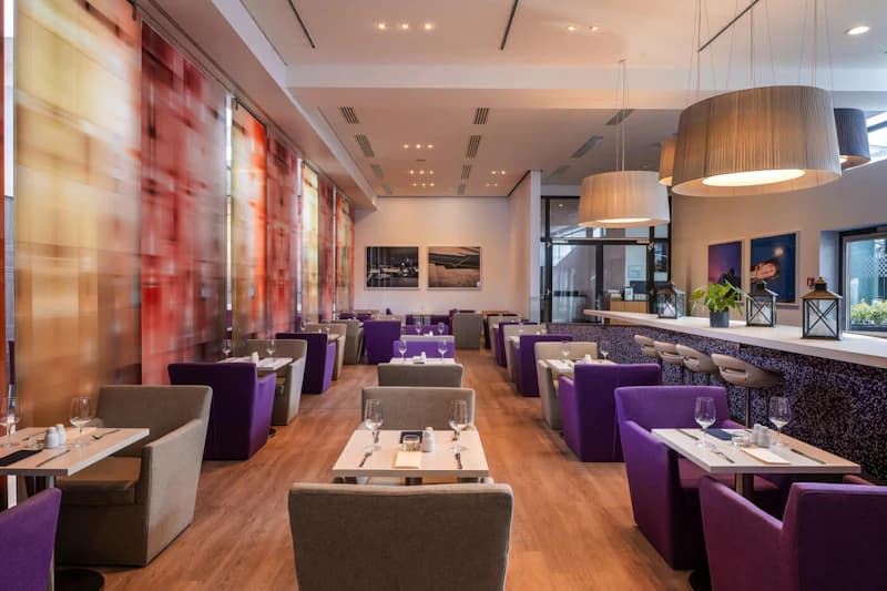 Hotelrestaurant mit Reihen gedeckter Tische, violetten Sesseln, Bartresen mit Hockern und großen Hängeleuchten.