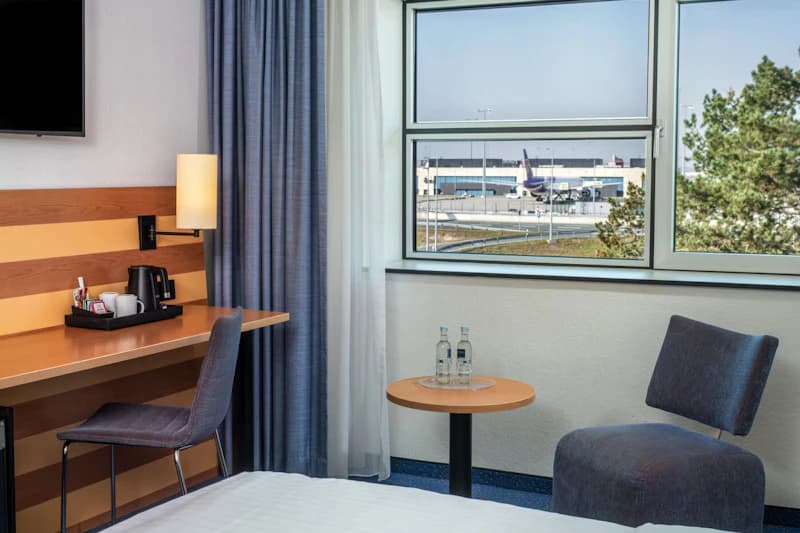 Hotelzimmer mit Schreibtisch, Kaffeemaschine, kleinem Tisch mit Wasserflaschen und Fensterblick auf Rollfeld und Flugzeug.