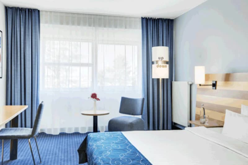 Helles Hotelzimmer mit Doppelbett, blauem Bettläufer, Fenster mit Vorhängen, Sitzgruppe mit kleinem Tisch und Schreibtisch.
