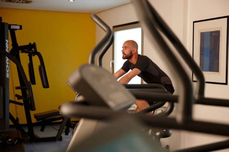 Mann auf Fahrrad-Ergometer in Hotelfitnessraum mit weiteren Trainingsgeraeten und gelber Wand.