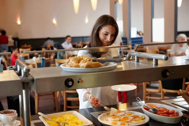 Frau steht am Frühstücksbuffet und nimmt ein Spiegelei, Croissants und Schalen mit Eierspeisen sichtbar.