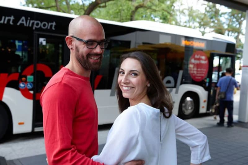 Paar umarmt sich vor einem Shuttlebus am Flughafenterminal, Mann mit rotem Pullover und Frau in weißer Bluse.