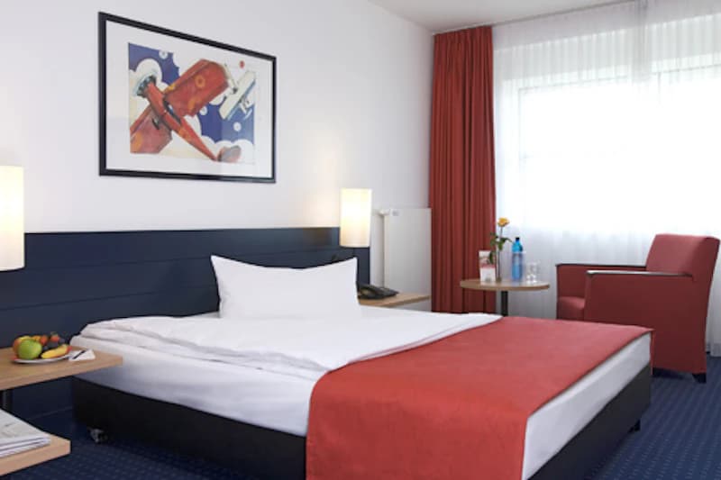 Hotelzimmer mit Doppelbett, rotem Bettläufer, Nachttischen, Sessel, kleinem Tisch mit Wasserflasche und Fenster mit Vorhang.