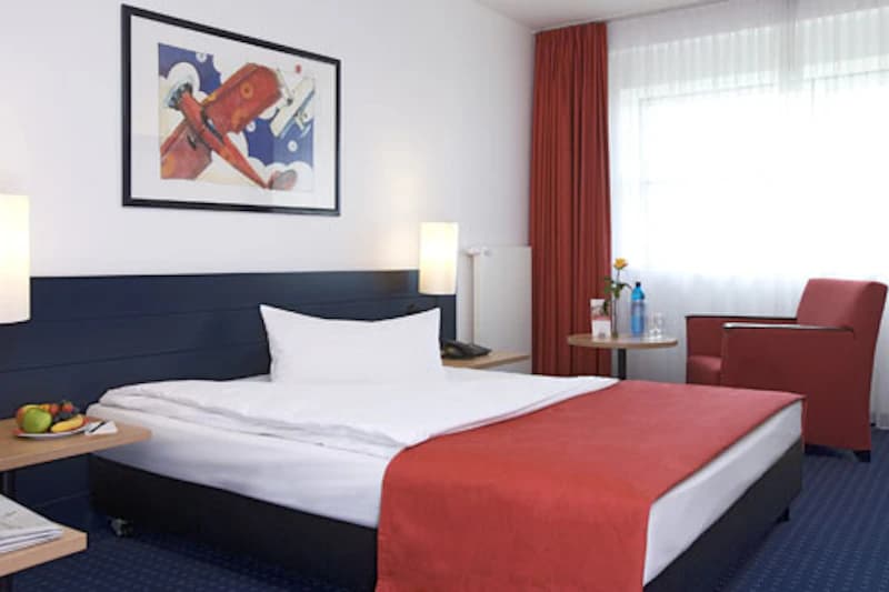 Hotelzimmer mit Doppelbett, roter Tagesdecke, Wandbild, Nachtlampen, Sessel und Beistelltisch.
