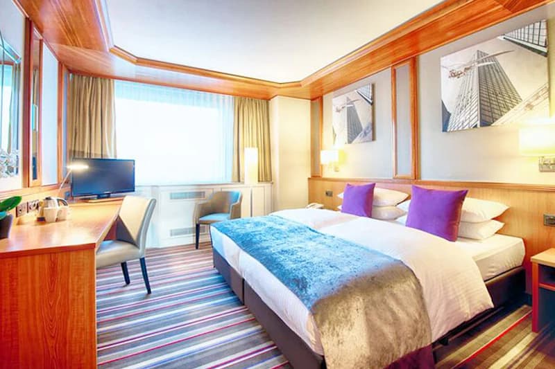 Hotelzimmer mit Doppelbett, lila Kissen, blauem Tagesdeckenläufer, Schreibtisch mit Fernseher und großem Fenster.
