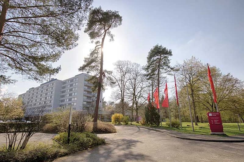 Mehrstöckiges Hotelgebäude links, Zufahrt mit Bäumen und mehreren roten Fahnen rechts, sonniger Himmel.