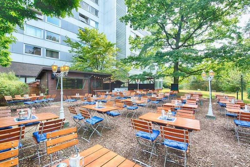 Außenbereich mit vielen Holztischen und blauen Sitzkissen unter Bäumen, Laternen und Hotelgebäude im Hintergrund.