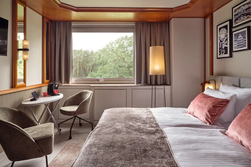 Hotelzimmer mit Doppelbett, Samtüberwurf, zwei Sesseln, kleinem Tisch mit Kaffeemaschine und Fenster mit Baumblick.