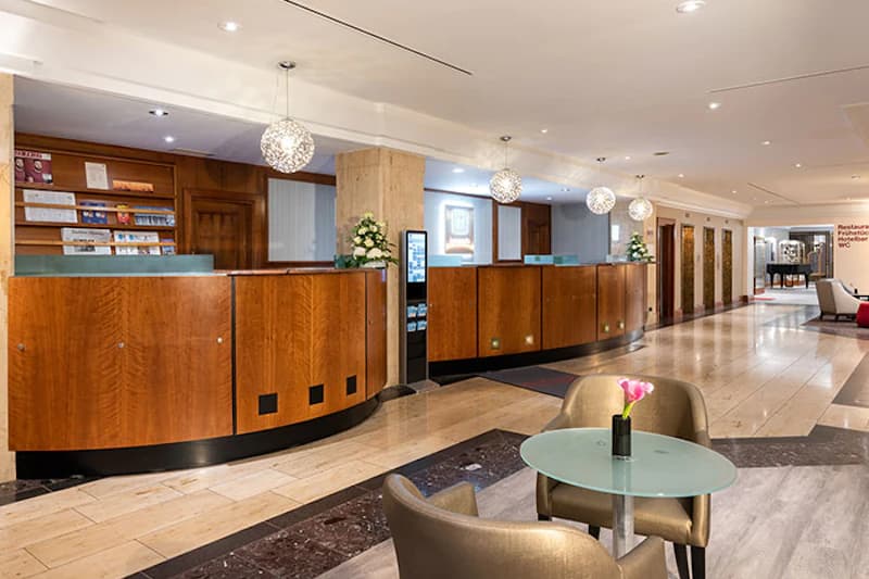 Große Hotelrezeption mit gebogenem Holztresen, Sitzgruppe mit Tisch und Blumen sowie hängenden Kugellampen.