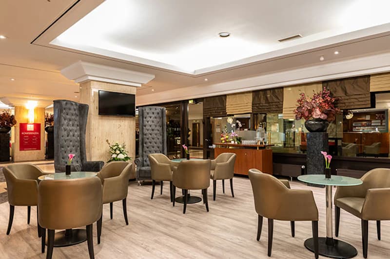 Hotellounge mit Tischen und braunen Stühlen, hohen grauen Sesseln, Fernseher an Säule und Blumendekor.
