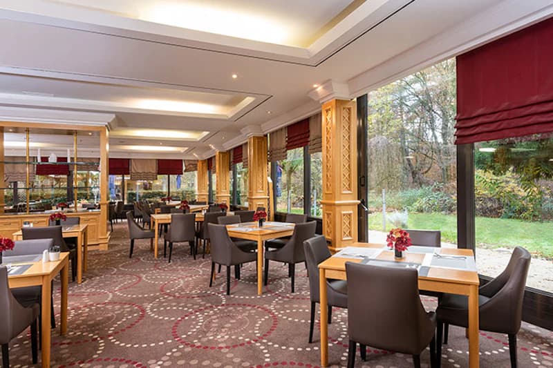 Helles Hotelrestaurant mit Holztischen, gepolsterten Stühlen und bodentiefen Fenstern zum Garten.