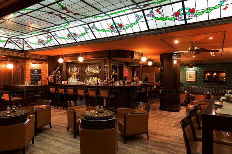 Innenraum einer Hotelbar mit hölzerner Theke, Barhockern, Ledersesseln und buntem Glasdach.
