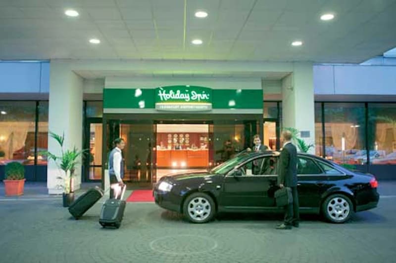 Hotelvorfahrt mit schwarzem Pkw und zwei Reisenden mit Koffern vor Eingang mit Holiday Inn Schriftzug.