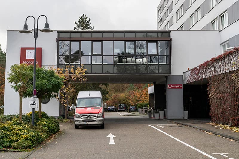 Hotelzufahrt mit überdachter Durchfahrt, rotes Lieferfahrzeug vor Gebäude mit Glasbrücke und Schild Rezeption.