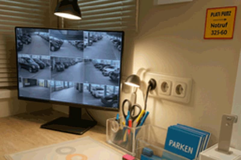 Überwachungstisch mit Bildschirm, der mehrere Parkhauskameras zeigt; Schreibtisch mit Stiftehalter und Parken-Broschüren.