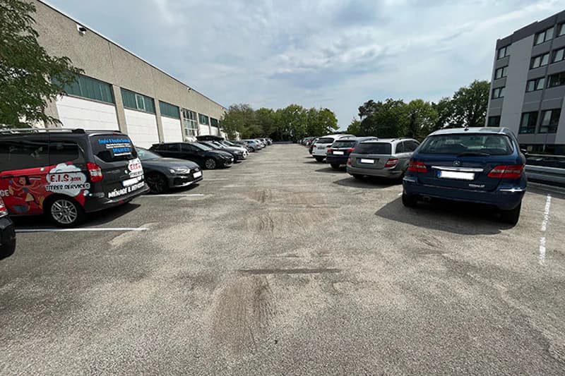 Asphaltierter Parkplatz mit Reihen geparkter Autos neben Industriehalle und Bürogebäude.