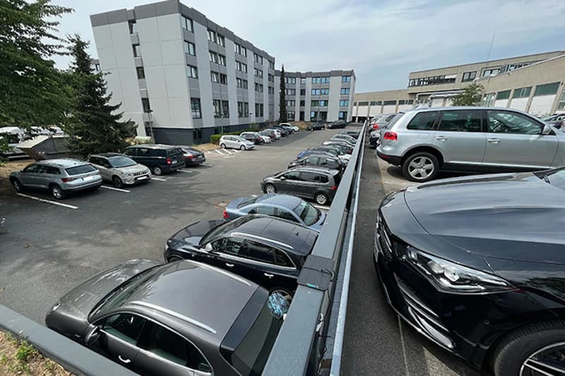 Zweistöckiger Parkplatz mit geparkten Autos, Metallgeländer und mehrstöckiges Bürogebäude im Hintergrund.