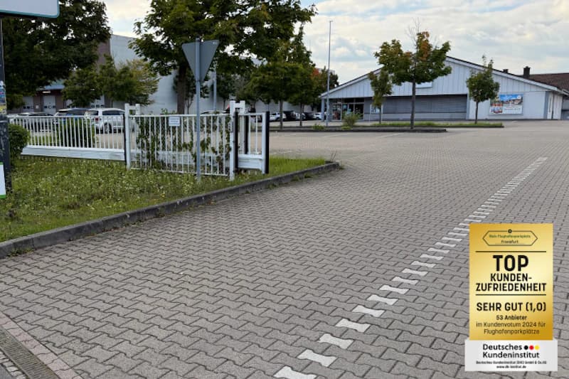 Parkplatz mit Pflasterbelag, geschlossenes Metalltor links, Gebäude und Bäume im Hintergrund, Auszeichnungstafel rechts.