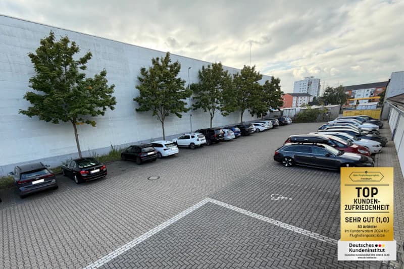 Gepflasterter Außenparkplatz mit Reihen geparkter Autos, Bäumen an weißer Wand und Auszeichnungstafel rechts unten.