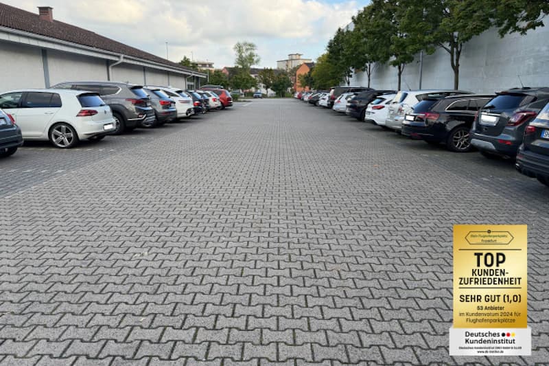 Großer Parkplatz mit geparkten Autos links und rechts auf gepflasterter Fläche und Bäumen im Hintergrund.