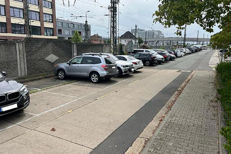 Reihen mit parkenden Autos entlang eines Parkplatzstreifens neben Bahntrasse und Gebäuden.