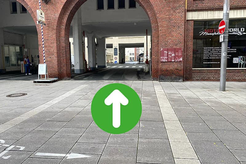 Pflasterplatz vor ziegelroten Torbögen mit Durchfahrt und grünem Kreis mit weißem Pfeil im Vordergrund.