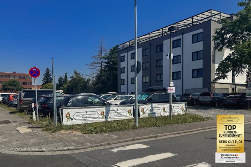 Freifläche mit geparkten Autos hinter niedriger Absperrung und Werbebannern, mehrstöckiges Gebäude im Hintergrund.