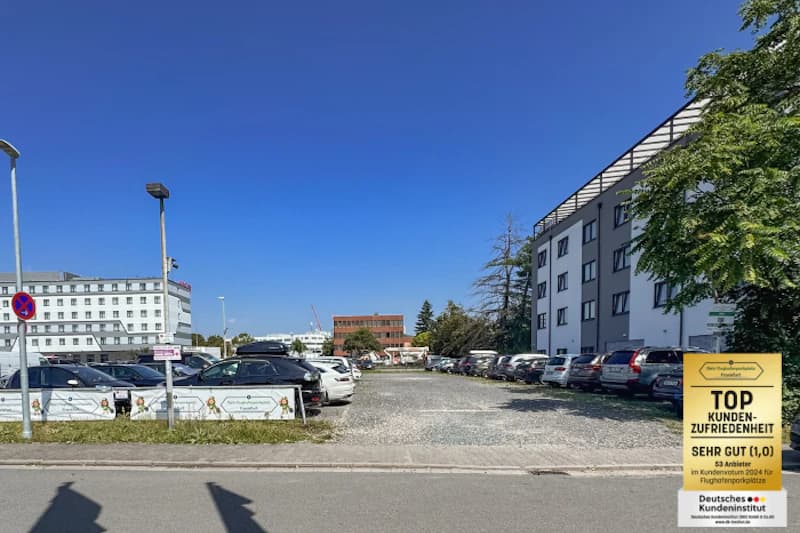 Schotterparkplatz mit geparkten Autos, Einzäunung mit Bannern und umliegenden Geschäftsgebäuden unter blauem Himmel.