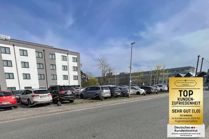 Freifläche mit geparkten Autos neben mehrgeschossigem Gebäude und gelbem Auszeichnungsschild am rechten Bildrand.