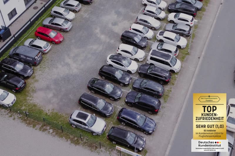 Luftaufnahme eines umzäunten Schotterparkplatzes mit Reihen geparkter Autos neben Gebäude und Straße.