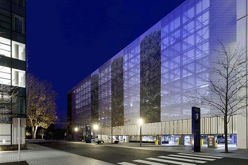 Mehrgeschossiges Parkhaus mit beleuchteter Fassade, offenen Einfahrten, Lampen und Bäumen bei Nacht.
