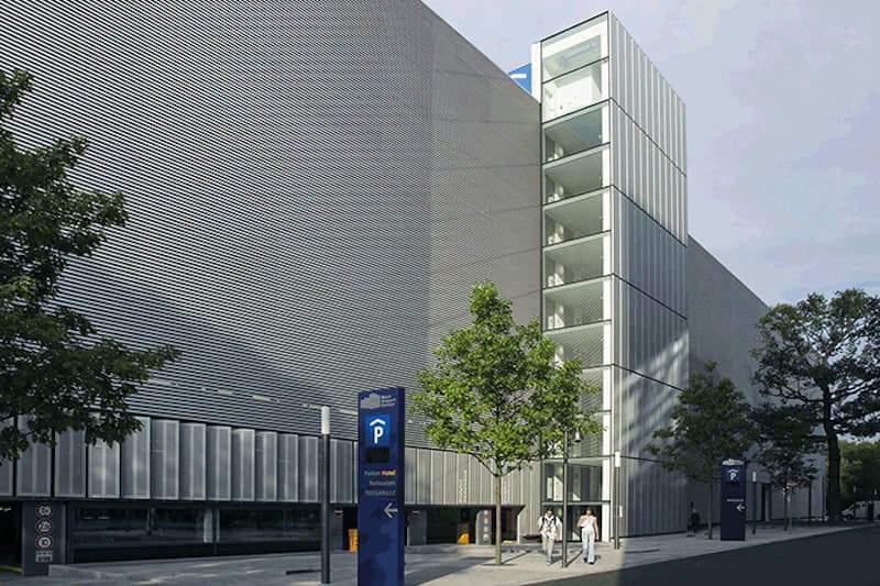 Mehrgeschossiges Parkhaus mit gläsernem Treppenturm, Parkhaus-Schildern, Bäumen und zwei Passanten.