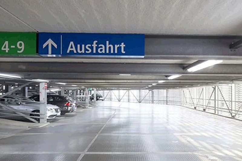 Mehrstöckiges Parkdeck mit geparkten Autos links, großem Ausfahrt-Schild an der Decke und beleuchtetem Fahrgang.