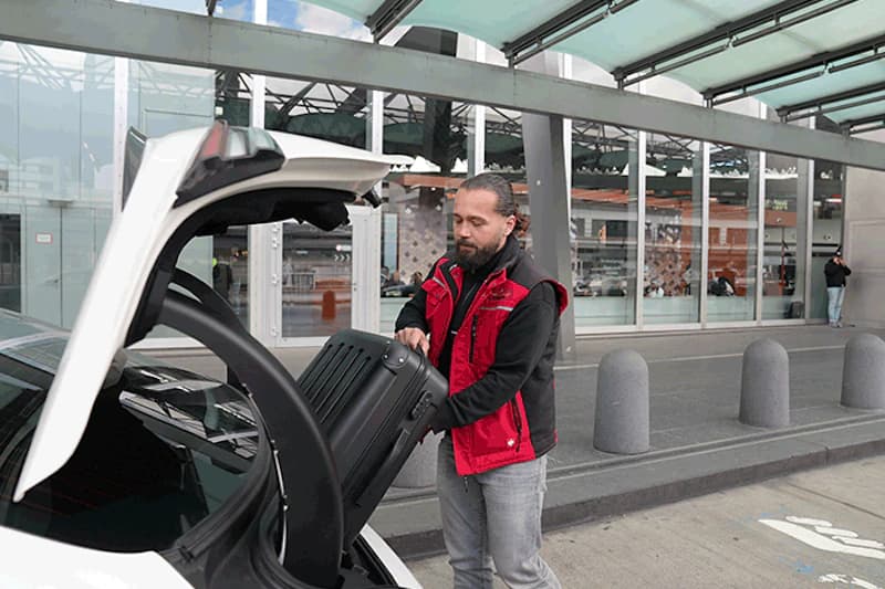 Mann in roter Weste lädt schwarzen Koffer in geöffneten Kofferraum vor Terminal.
