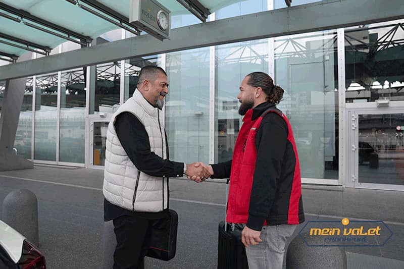 Zwei Männer schütteln sich die Hand vor verglastem Flughafen-Terminal, beide mit Reisekoffern, einer trägt rote Weste mit Mein-Valet-Logo.