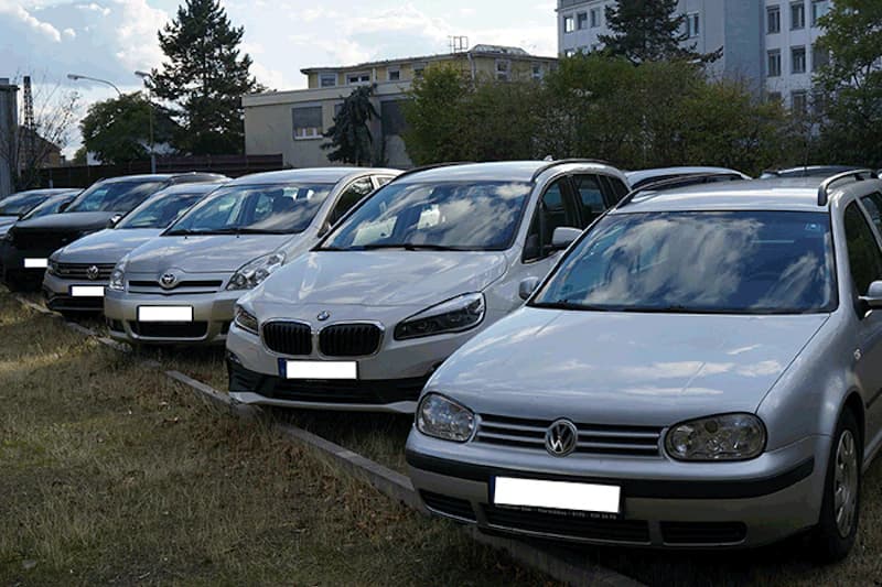 Mehrere geparkte Pkw in einer Reihe auf einem Außenstellplatz neben einem Grasstreifen, Gebäude im Hintergrund.