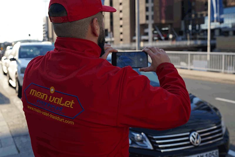 Mitarbeiter in roter Mein-Valet-Jacke fotografiert mit Smartphone die Front eines schwarzen Volkswagen am Straßenrand.
