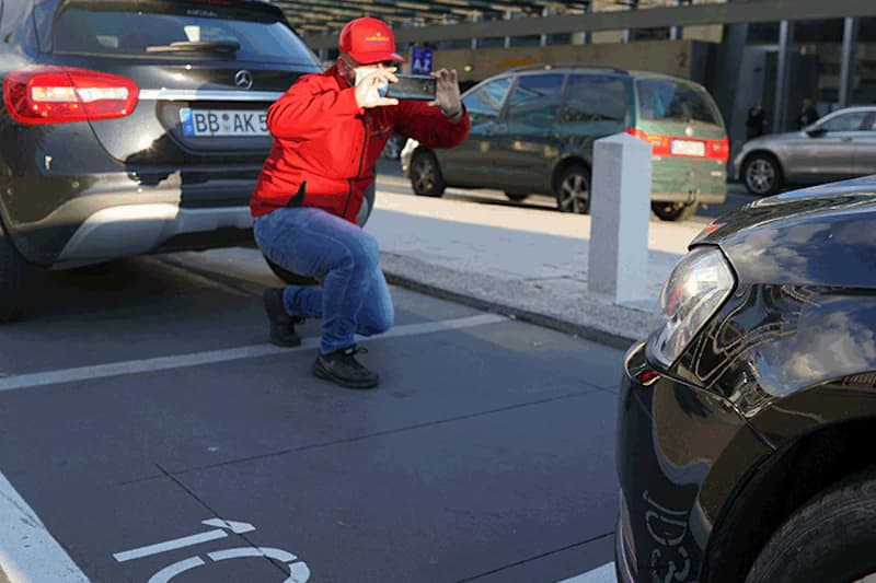 Person in roter Jacke kniend fotografiert ein geparktes Auto mit Smartphone auf Parkplatz am Flughafen Frankfurt.