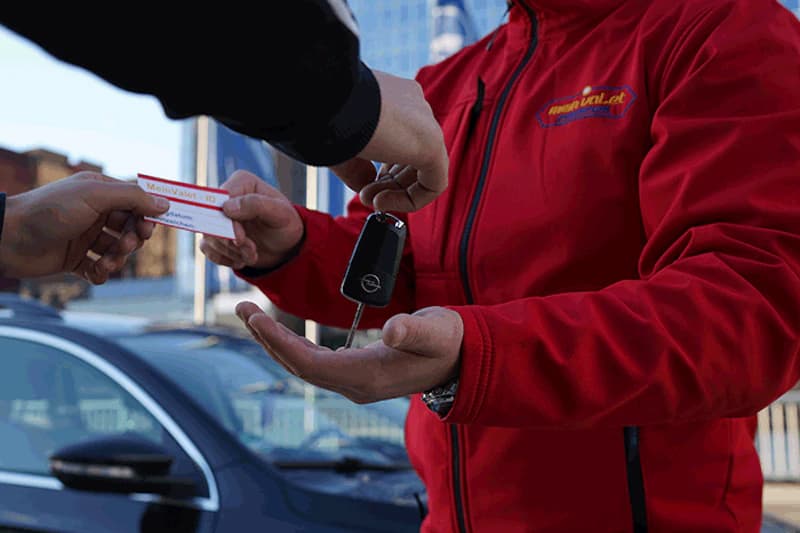 Mitarbeiter in roter Jacke übergibt Autoschlüssel und Abholschein vor geparktem Auto.