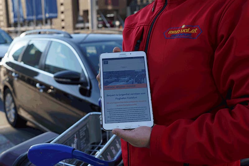 Person mit roter Mein-Valet Jacke hält ein Tablet vor einem geparkten Auto und Einkaufswagen.