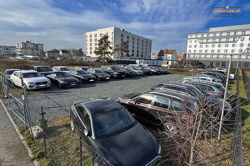 Umzäunter Schotterparkplatz mit mehreren Reihen geparkter Autos und Wohngebäuden im Hintergrund.