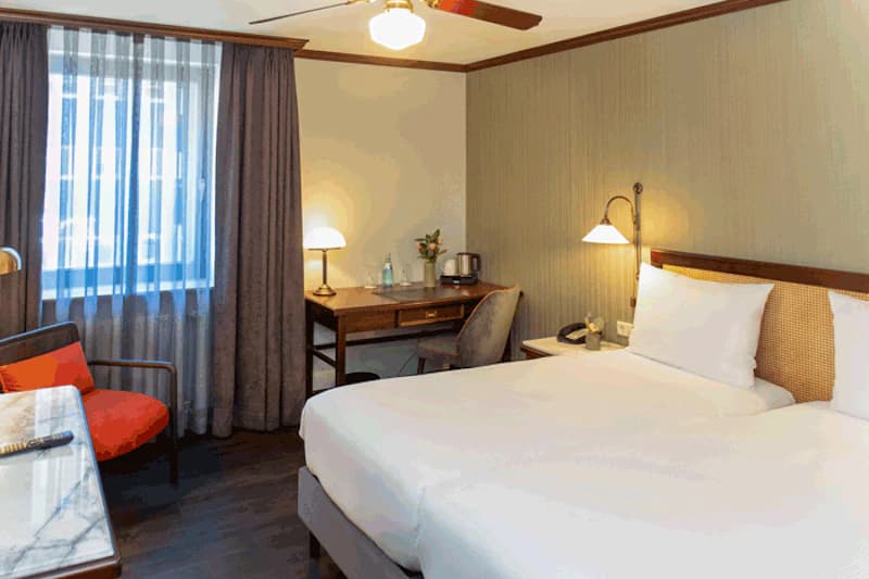 Helles Hotelzimmer mit zwei Einzelbetten, Nachttischlampen, Schreibtisch, Sessel und Gardinen.