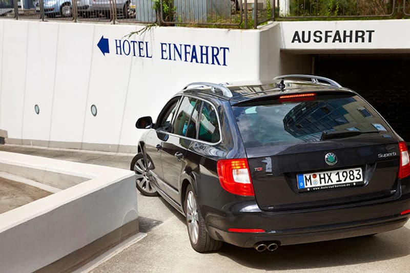 Dunkler Kombi fährt in eine steile Hotelgarageneinfahrt vor Wand mit Schrift Hotel Einfahrt und Ausfahrt.