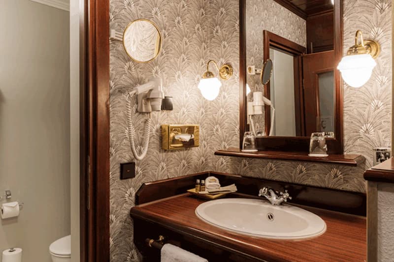Hotelbad mit Holzwaschtisch, rundem Waschbecken, Wandspiegel, Haartrockner und gemusterter Tapete; WC teilweise sichtbar.